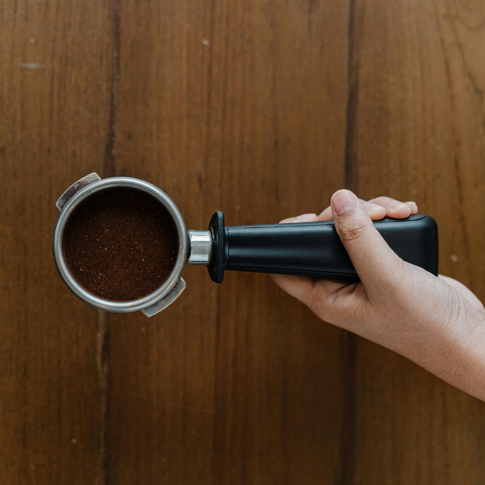 A birds eye view image of a hand holding a portafilter, perfectly centred over a wooden backdrop. The coffee grounds have been tamped.