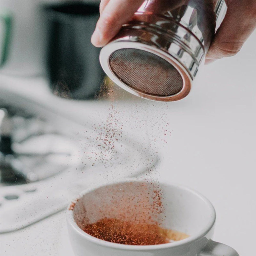 A close up image of a hand tipping cocoa out of a shaker. The cocoa is being dusted into an empty white coffee mug